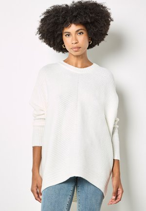 Przesadnie duży biały sweter z dzianiny o prążkowanej fakturze, okrągłym dekoltem i opuszczanymi rękawami, zestawiony z jasnoniebieskimi dżinsami.