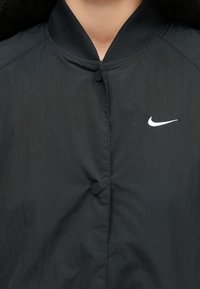 Μαύρο μπουφάν τύπου bomber με ομαλή υφή, ριμπ γιακά και δύο κυκλικά κουμπιά. Διαθέτει λευκό λογότυπο Nike στην αριστερή πλευρά.