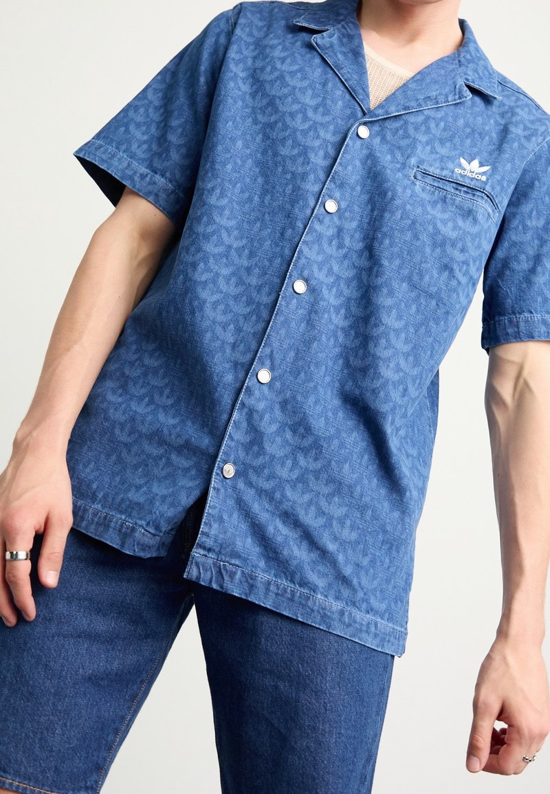 Chemise à manches courtes, bleue, à boutons avec un motif texturé, détail de poche blanche et marquage. Fabriquée en tissu léger.