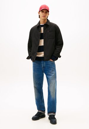 Schwarze Knopfjacke über einem gestreiften Pullover, kombiniert mit blauen Jeans und schwarzen Sneakers. Die rote Kappe rundet das lässige Outfit ab.