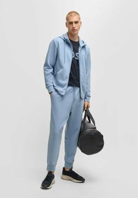 Ljusblå zip-up hoodie, matchande joggers, svart t-shirt, svarta sneakers och en rund svart väska. Slät tyg, avslappnad passform och minimalistisk design.