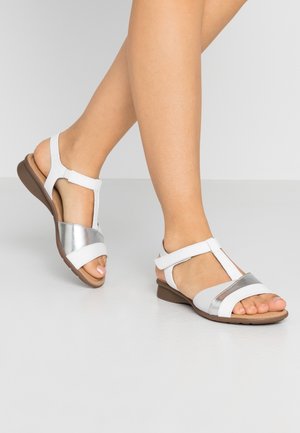 White and silver sandals with a T-strap design, featuring a flat sole and soft material. Adjustable ankle strap for secure fit.