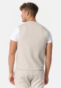 Chaleco sin mangas de color beige claro, hecho de una tela suave, que presenta un diseño sencillo con dos costuras traseras, llevado sobre una camiseta blanca.