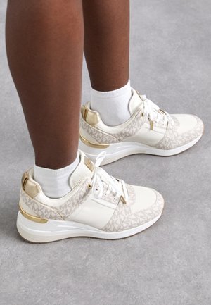 White sneakers with beige patterned textile upper, gold accents, and a white rubber sole. Features classic lacing and cushioned ankle support.