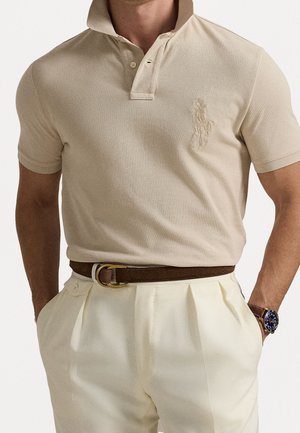Homme portant un polo beige rentré dans un pantalon plissé crème avec une ceinture marron et une montre-bracelet avec un cadran bleu.