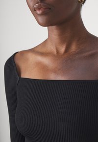 Haut à manches longues noir à côtes avec un décolleté carré. Le tissu a une texture lisse, mettant en valeur le design ajusté sur les épaules.