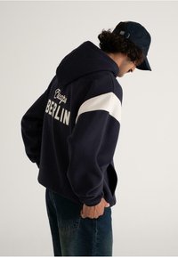 Man wearing navy hoodie with white stripe and "Escape Berlin" text on back, navy cap, and blue jeans, standing against plain background.