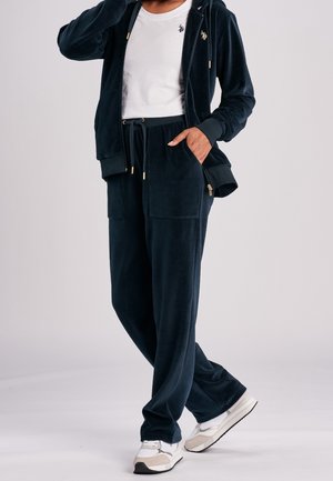 Marineblauw velours trainingspak met capuchon en ritsjack en broek met trekkoord. Voorzien van grote zakken en gouden details. Gecombineerd met witte sneakers.