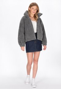 Šedá huňatá bunda s vysokým límcem, zipem a oversized rukávy, v kombinaci s džínovou mini sukní a bílými teniskami.
