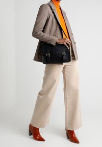 Borsa nera con due fibbie frontali, abbinata a un blazer a scacchi, maglione a collo alto arancione, pantaloni larghi crema e décolleté rossi con punta.