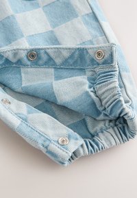 Lyseblåt jeansstof med ternet mønster. Har metalknapper og elastik samlet ved manchetterne. Blød tekstur.