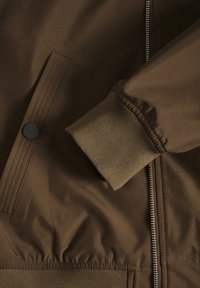 Gros plan de la manche d'une veste marron avec poignet côtelé, fermeture éclair et poche à pression sur une surface en tissu lisse.
