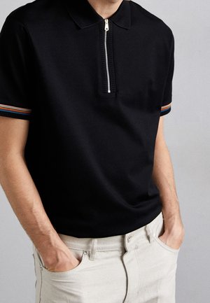 Man wearing black short-sleeve polo shirt with zipper and striped sleeve cuffs, hands in pockets of light beige pants, neutral background.