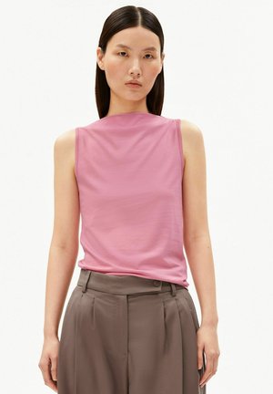 Jeune femme aux cheveux lisses et foncés porte un haut rose sans manches et un pantalon taupe taille haute, debout devant un fond blanc uni.