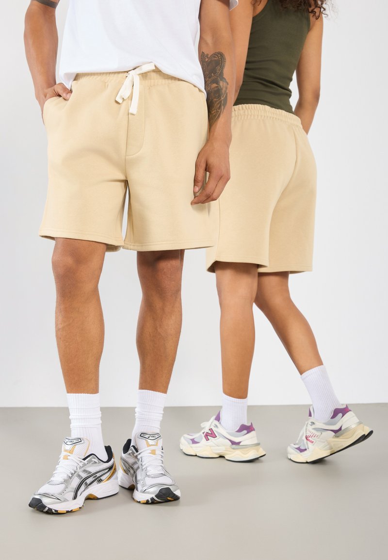 Shorts en coton beige avec taille élastique et cordon de serrage, associés à des baskets blanches et grises. Design simple, texture lisse.