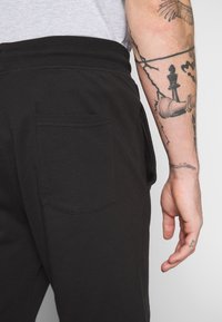 Pantalones de chándal negros con una cinturilla acanalada y un solo bolsillo trasero, fabricados en un tejido suave. Tatuaje visible en el brazo que sostiene los pantalones.