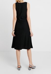 Schwarzes geripptes ärmelloses Kleid mit einem ausgestellten Saum. Getragen mit schwarz-weißen gemusterten Schuhen mit Blockabsatz und Knöchelriemen.