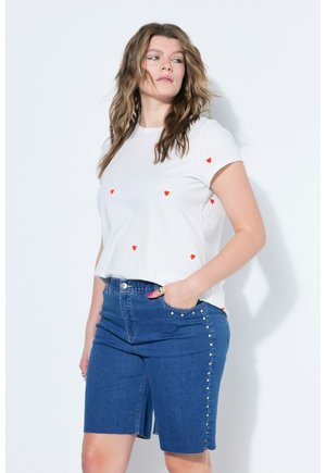 Frau mit welligem Haar, die ein weißes T-Shirt mit kleinen roten Herzen und knielange blaue Jeansshorts mit Perlenakzenten an den Seiten trägt.