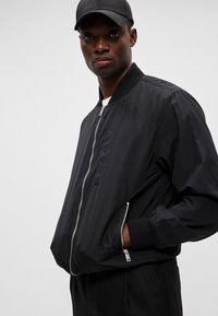 Schwarze Bomberjacke aus glattem Material, mit einem Reißverschluss auf der Vorderseite, Seitentaschen, gerippten Bündchen und Kragen, im klassischen Schnitt.