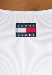 Vit ribbad tyg med en marinblå rektangulär lapp som har "TOMMY" i vitt och "JEANS" nedan i vitt, accentuerad med röda och vita ränder.