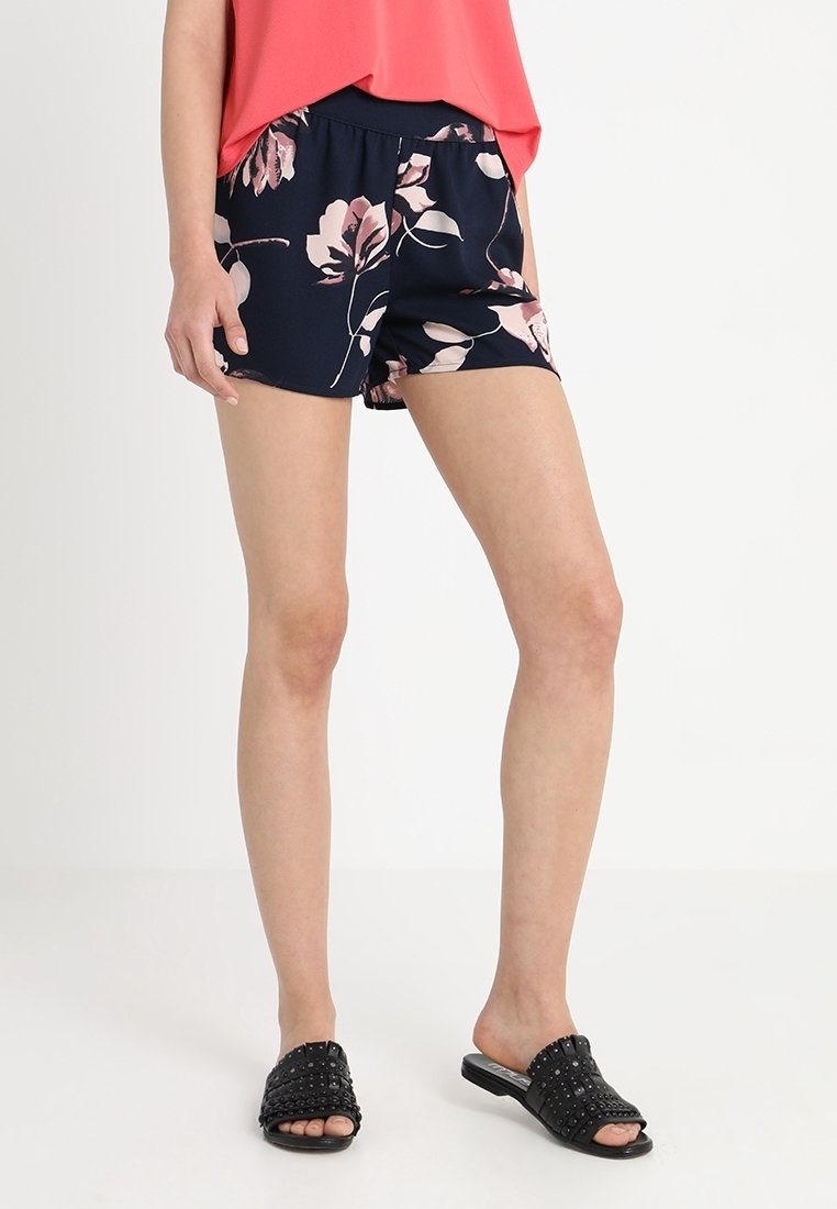 Woman wearing navy blue floral shorts, coral sleeveless top, and black studded slip-on sandals, standing against a white background.