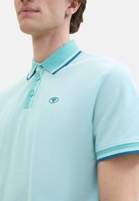 Lichtblauwe polo met een gestructureerde stof, voorzien van een twee-knopenknopsluiting en contrasterende donkerblauwe strepen op de kraag en mouwen.