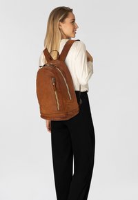 Sac à dos en cuir marron avec une texture lisse, des compartiments zippés et des bretelles ajustables. Il présente un fond arrondi et un design élégant.