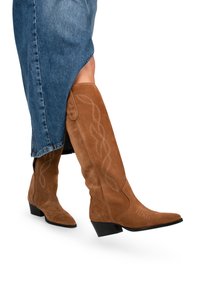 Botas altas de ante marrón de estilo western, con punta afilada, que presentan costuras decorativas y un tacón bajo tipo bloque. Se llevan con una falda de mezclilla.