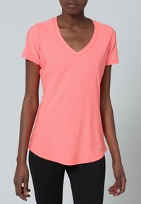 Mujer vestida con una camiseta de cuello en V de color rosa coral y leggings negros, de pie contra un fondo gris neutro.