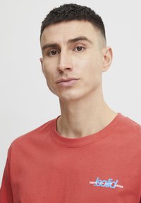 Rød bomulls t-skjorte med rund hals og korte ermer, med et blått og hvitt grafisk trykk som sier "Tokyo" og "solid Japan."