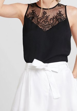 Femme portant un haut noir sans manches avec un détail en dentelle et un short blanc taille haute noué par un nœud à la taille.