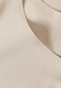 Beige stof met een zachte textuur, voorzien van een ronde halslijn en gelaagde secties. Het materiaal lijkt lichtgewicht en zacht te zijn.