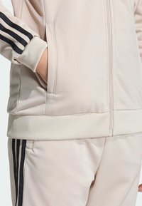 Giacca beige con cerniera, polsini a coste, tasche laterali e dettagli neri a tre strisce sulle maniche. Tessuto liscio ed elastico.