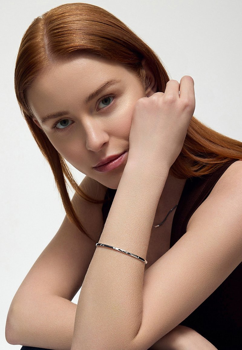 Srebrna bransoletka o skręconym wzorze na nadgarstku modelki. Posiada gładkie, lśniące powierzchnie i elegancki, minimalistyczny wygląd.