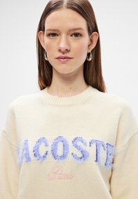 Κρεμ φόρεμα με απαλή υφή. Διαθέτει φουντωτό ανοιχτό μπλε κέντημα "LACOSTE" και ροζ διακόσμηση "Παρίσι". Στρογγυλή λαιμόκοψη.