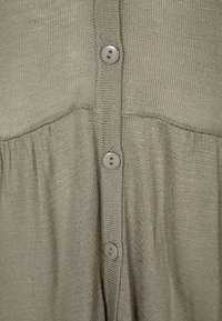 Indumento lavorato a maglia verde chiaro con finitura testurizzata, caratterizzato da sei bottoni tondi di colore neutro sul davanti e un dettaglio arricciato in vita.