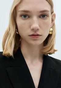 Blonde Haare mit sanften Wellen, tragt goldene, runde Ohrringe. Gekleidet in einem schwarzen Blazer mit spitzen Revers, minimalem Muster, klare Haut.