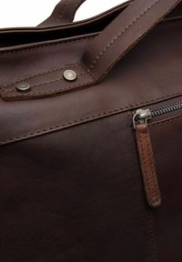 Sac en cuir marron avec une poche zippée et des accents en métal. Texture lisse avec des coutures détaillées et des rivets.