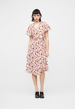 Γυναίκα που φοράει λευκό φόρεμα τύπου wrap με κόκκινο και μπλε λουλουδένιο σχέδιο και μανίκια με βολάν, συνδυασμένο με μαύρα παπούτσια με τετράγωνη μύτη, στέκεται μπροστά σε λευκό φόντο.