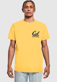 Žluté tričko s krátkým rukávem a kulatým výstřihem, které je zdobeno námořnickým logem s nápisy "Cal" a "University of California" na levé straně hrudníku.