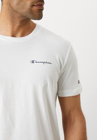 White cotton t-shirt featuring a navy "Champion" logo on the chest and a small logo on the sleeve. Short sleeves with a round neckline.