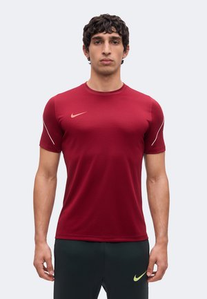 Mężczyzna w czerwonej koszulce Nike z krótkim rękawem i ciemnych spodniach sportowych z żółtym logo Nike, stojący na jednolitym jasnym tle.