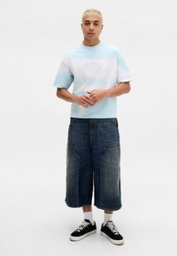 Jonge persoon met kort blond haar die een lichtblauw gestreept shirt draagt, brede denim knielange shorts, witte sokken en zwarte sneakers, staand.