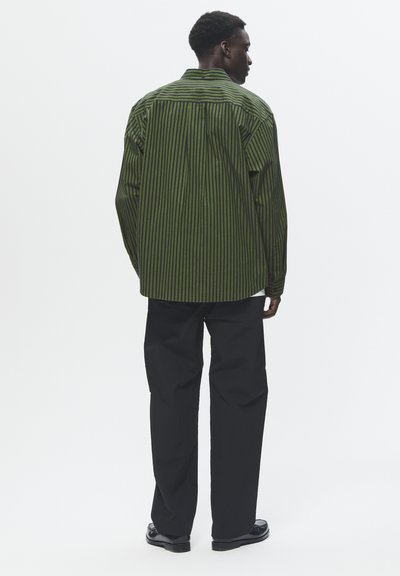 Green and black striped shirt with a relaxed fit, long sleeves, and a curved hem. Paired with black pants and black shoes.