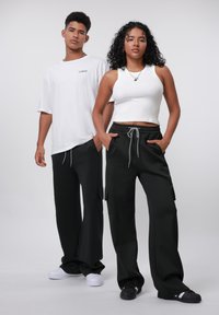 Twee modellen staan naast elkaar. Het mannelijke model draagt een losse witte t-shirt en zwarte broek. Het vrouwelijke model heeft een strakke witte tanktop aan en zwarte broek met zakken. Beiden dragen casual schoenen.