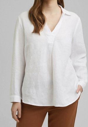 Kvinde iført en løs hvid langærmet V-hals bluse med krave, kombineret med brune bukser, stående foran en ensfarvet baggrund.