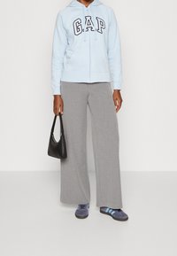 Persona con una sudadera con cremallera de GAP en azul claro, pantalones anchos grises, zapatillas deportivas azules, sosteniendo un pequeño bolso negro a un lado.