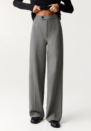 Personne portant un pantalon ample gris taille haute avec boutons sur le devant, chaussures noires et haut noir relevé pour dévoiler le ventre.