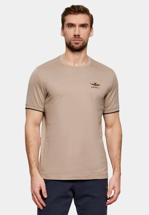 Uomo con capelli castani corti che indossa una maglietta beige a maniche corte con scollo rotondo e un piccolo emblema sul petto sinistro, in piedi davanti a uno sfondo semplice.