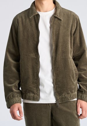 Mann trägt olivgrüne Cord-Strickjacke mit Reißverschluss und passende Hose über einem schlichten weißen T-Shirt mit Rundhalsausschnitt.